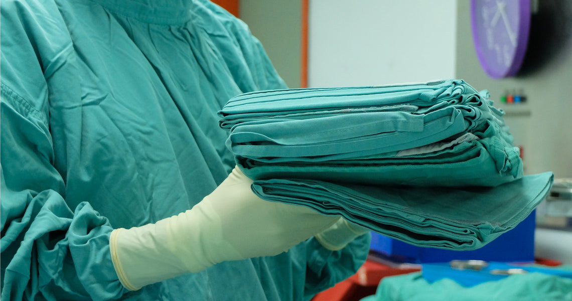 Person in surgical scrubs and gloves holding a stack of folded green medical drapes in a clinical setting.