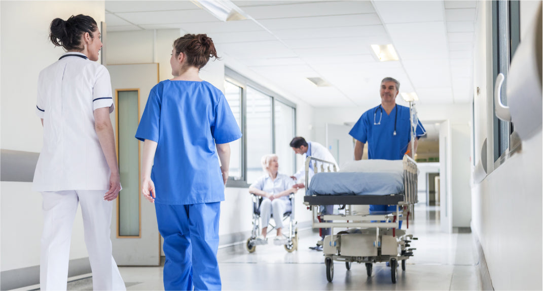 Healthcare professionals and staff move a patient in a wheelchair and an empty gurney through a brightly lit hospital corridor. Two nurses are talking in the foreground.