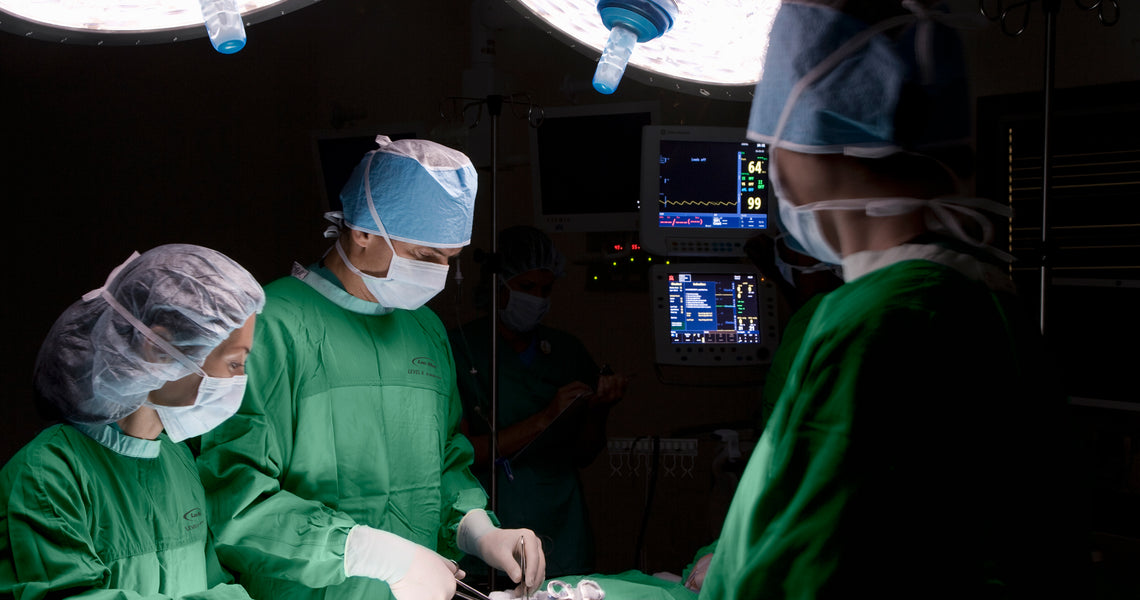 Two surgeons in a dimly lit operating room perform a surgery under a bright overhead light, with medical equipment and monitors in the background.